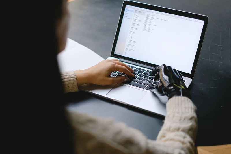A person’s hand and a robotic hand typing together on the same laptop